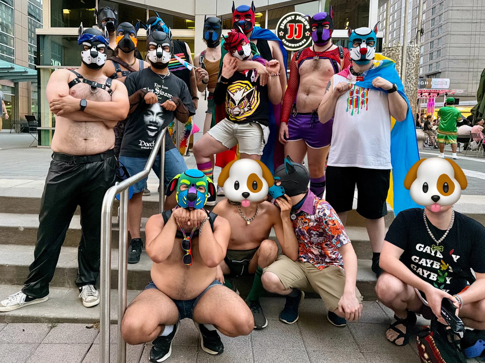 A group of people, many wearing stylized leather and neoprene puppy hood masks, gather for a photo on outdoor concrete steps during a Pride event. In the foreground, one person in a bright green and blue puppy hood crouches forward, while next to them, a shirtless individual playfully leans in toward another person wearing a black and blue puppy hood and a colorful patterned shirt. Other attendees stand on the steps behind them, wearing various gear, harnesses, rainbow capes, and Pride-themed apparel. A Jimmy John's sign is visible in the background, and a "High Pride" stage banner can be seen in the distance to the right.