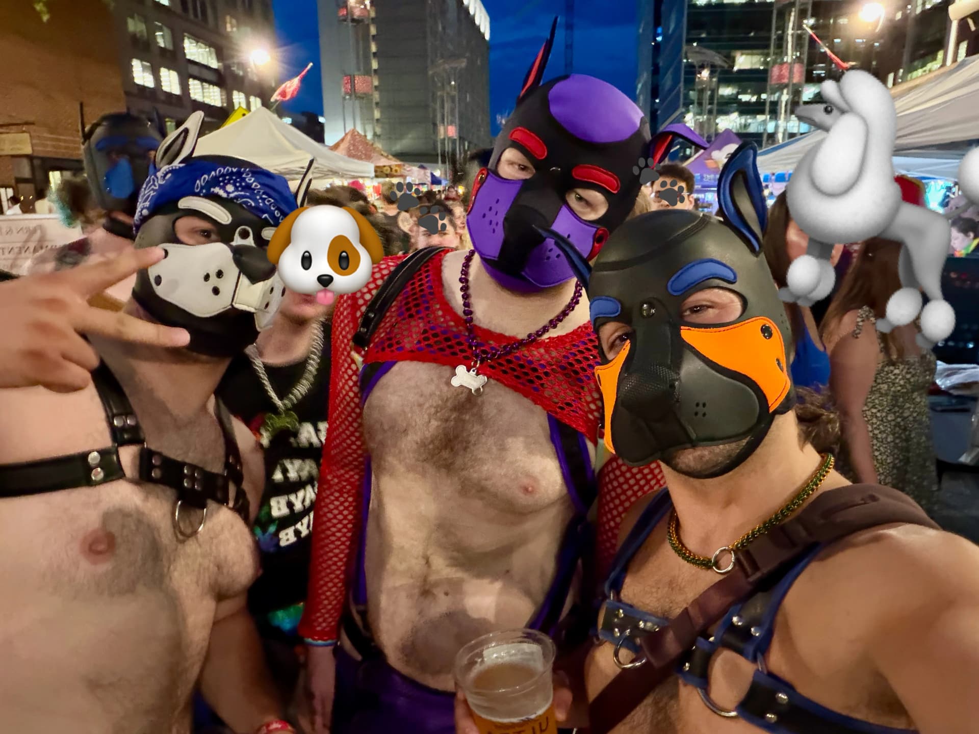 A close-up group selfie taken at dusk during an outdoor street festival or market. In the foreground, three individuals wear stylized leather and neoprene puppy hood masks and body harnesses. The person on the far left wears a blue bandana over a black-and-white mask, making a peace sign at the camera. The person in the center wears a purple and black mask, a red fishnet crop top, and holds a clear plastic cup of beer. The individual on the right, taking the selfie, wears a black mask with distinct blue and orange markings. In the blurred background, other festival attendees, white vendor tents, and city buildings are visible under a twilight sky.